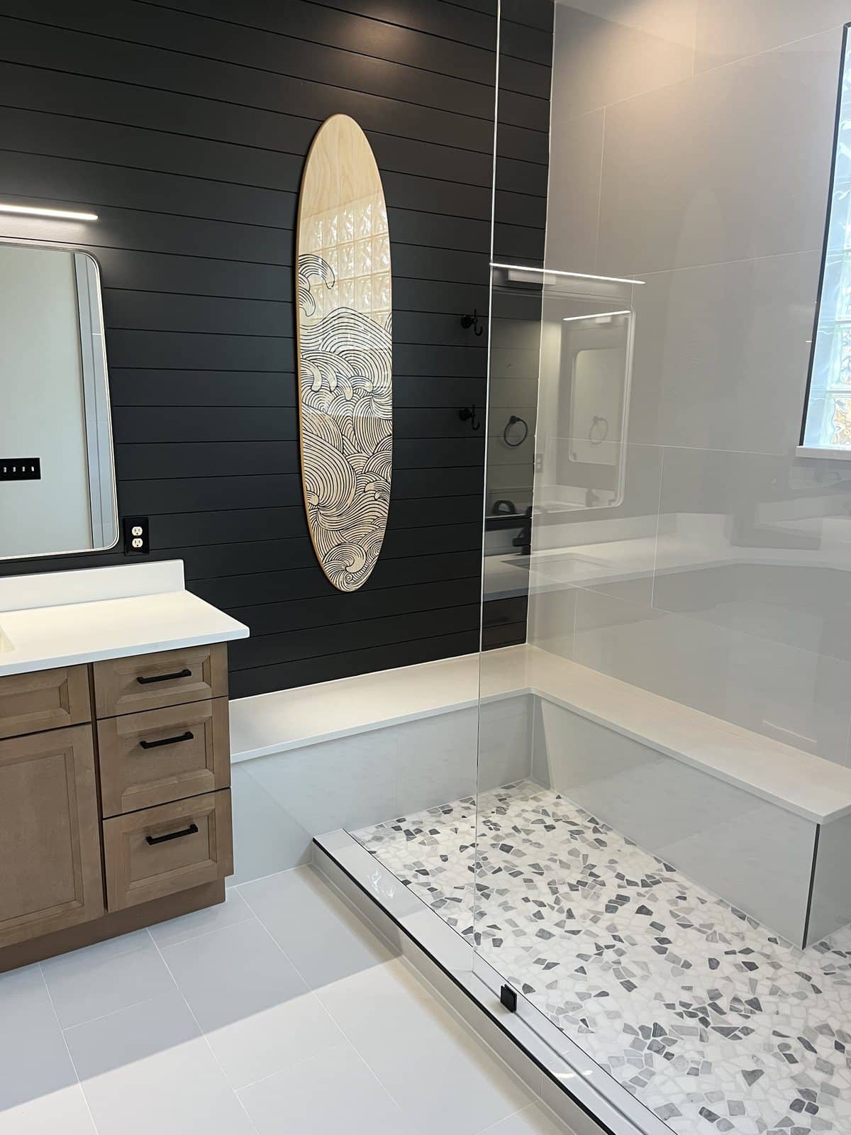 Bathroom with a black shiplap wall, a surfboard mounted as art, a floating wood vanity, a curbless glass shower, and a pebble floor.