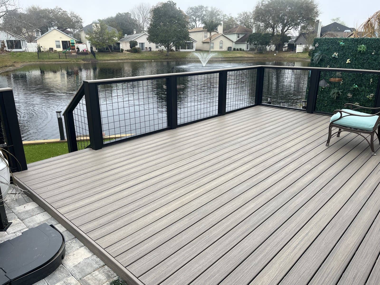 Composite deck with black metal posts and hog-wire railing overlooking a pond, with a covered seating area and a wooded far shore.