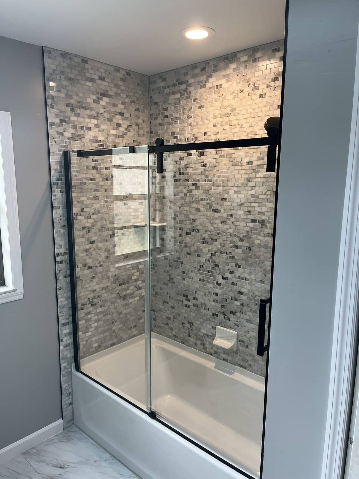 Soaking tub in a marble mosaic tile surround with a black-framed sliding glass door, a rainfall shower head, and a glass niche.